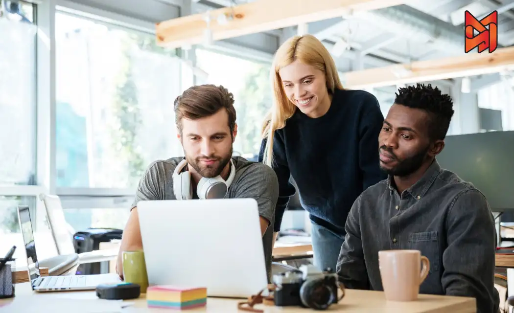 Three digital marketing team members brainstorm a social media ad campaign, gathered around a laptop analyzing analytics dashboards